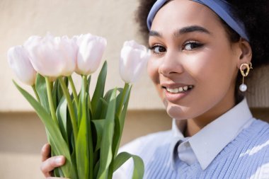 close up of cheerful african american woman in blue vest and white shirt holding bouquet of tulips near wall
