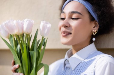 cheerful african american woman in blue vest and white shirt looking at tulips near wall