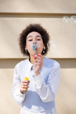 Mavi yelekli ve beyaz gömlekli genç Afro-Amerikan kadın sabun köpüğü üflüyor.