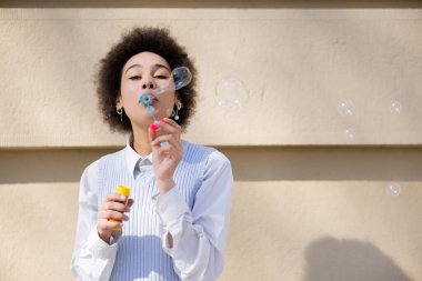 curly african american woman holding bottle and blowing soap bubbles near wall