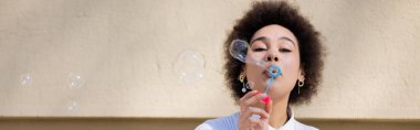 curly african american woman blowing soap bubble near wall, banner