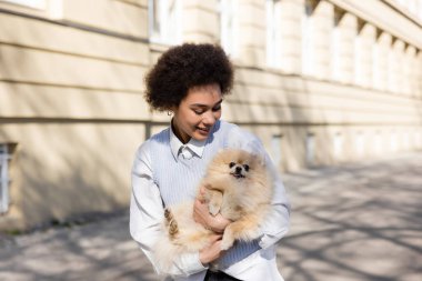 cheerful african american woman holding pomeranian dog on street