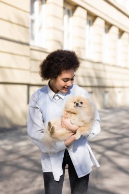 cheerful african american woman in blue vest holding pomeranian dog on street