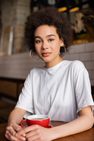 pleased african american woman in hoop earrings looking at camera near cup of coffee 