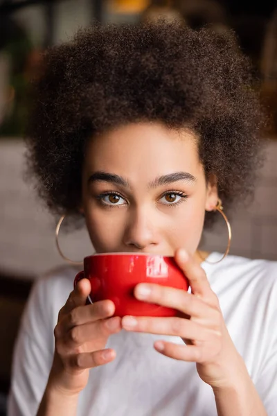 african american woman in hoop earrings drinking coffee and looking at camera