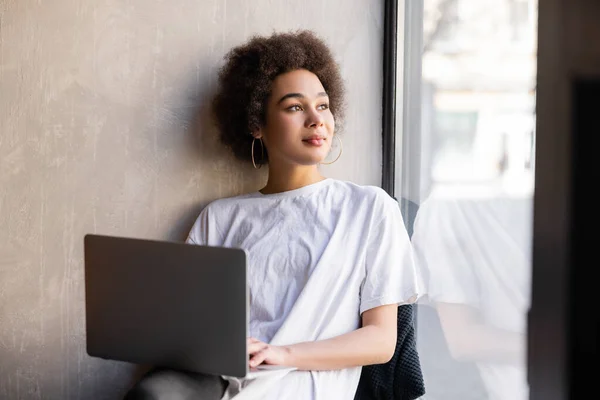 young african american woman in white t-shirt holding laptop near window 