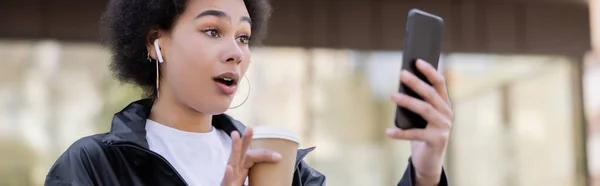 shocked african american woman in earphone holding paper cup and having video chat on smartphone, banner