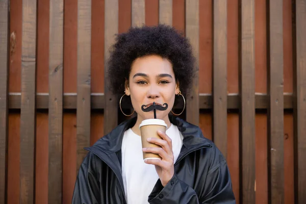 curly african american woman holding paper cup and straw with moustache near fence