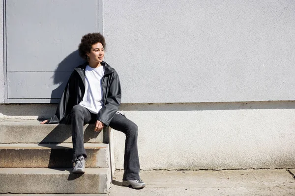 full length of happy african american woman in jacket sitting on stairs near wall
