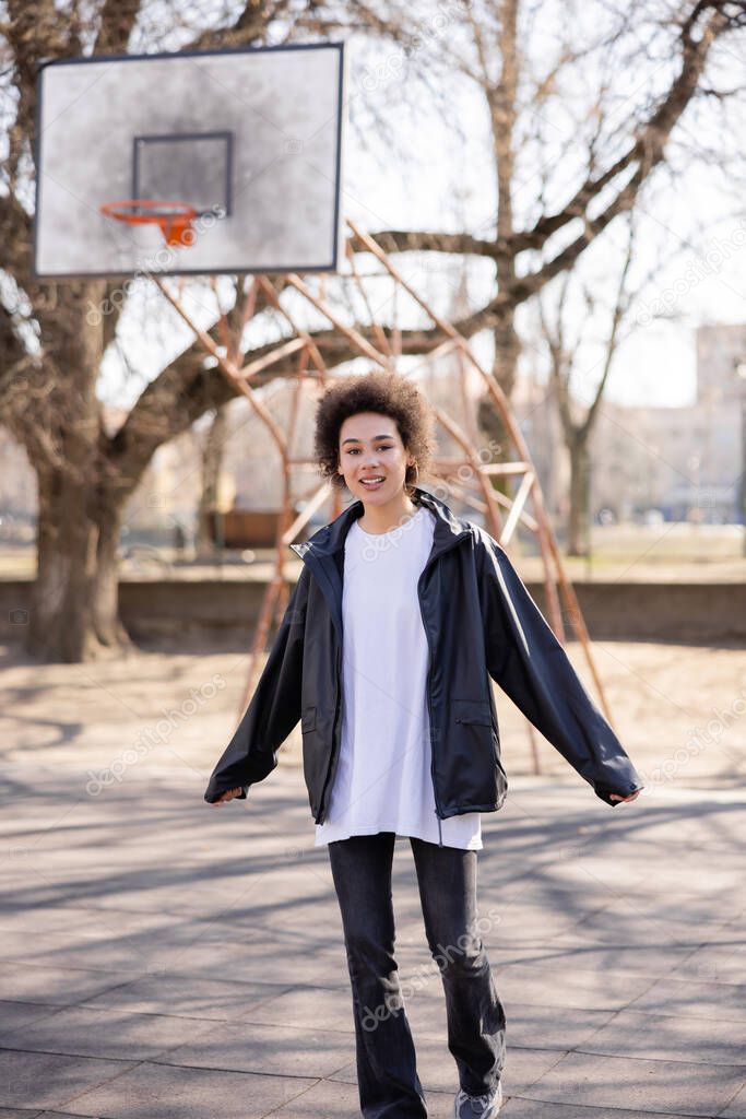 happy african american woman in jacket on basketball outdoor court