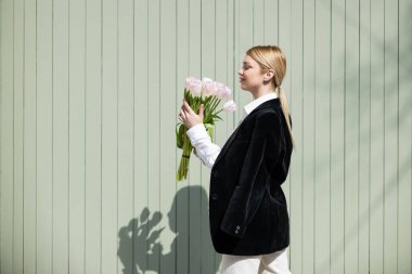 Siyah ceketli genç bir kadın gri duvarın yanında beyaz laleler tutuyor.