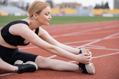 Genç sporcu kadın stadyumda otururken bacağını esnetiyor.