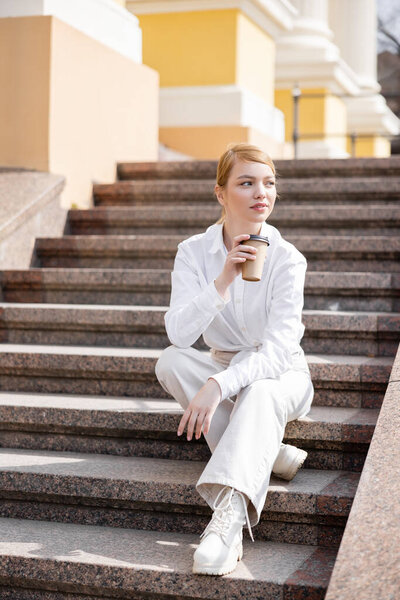 full length view of woman in white clothes sitting on stairs with coffee to go
