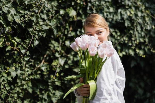 happy young woman obscuring face with white tulips near green ivy