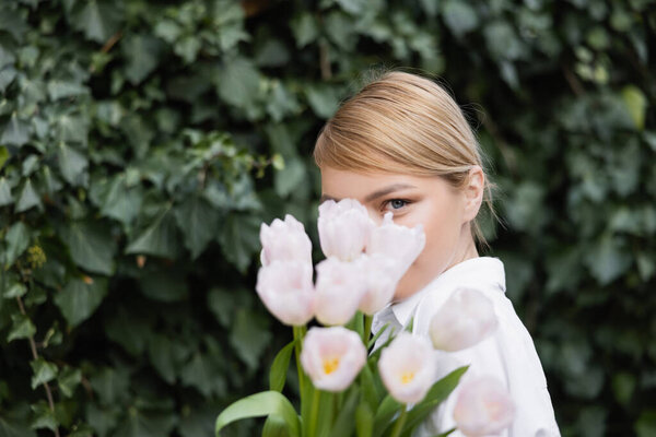 blonde woman obscuring face with white tulips and looking at camera 