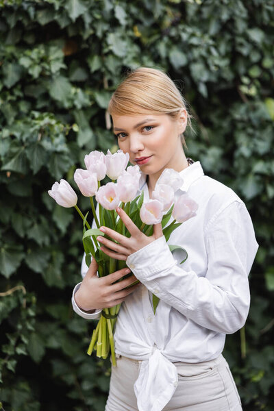 blonde woman in white shirt holding white tulips and looking at camera near green ivy