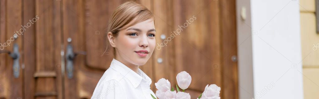 blonde young woman smiling near fresh tulips outdoors, banner