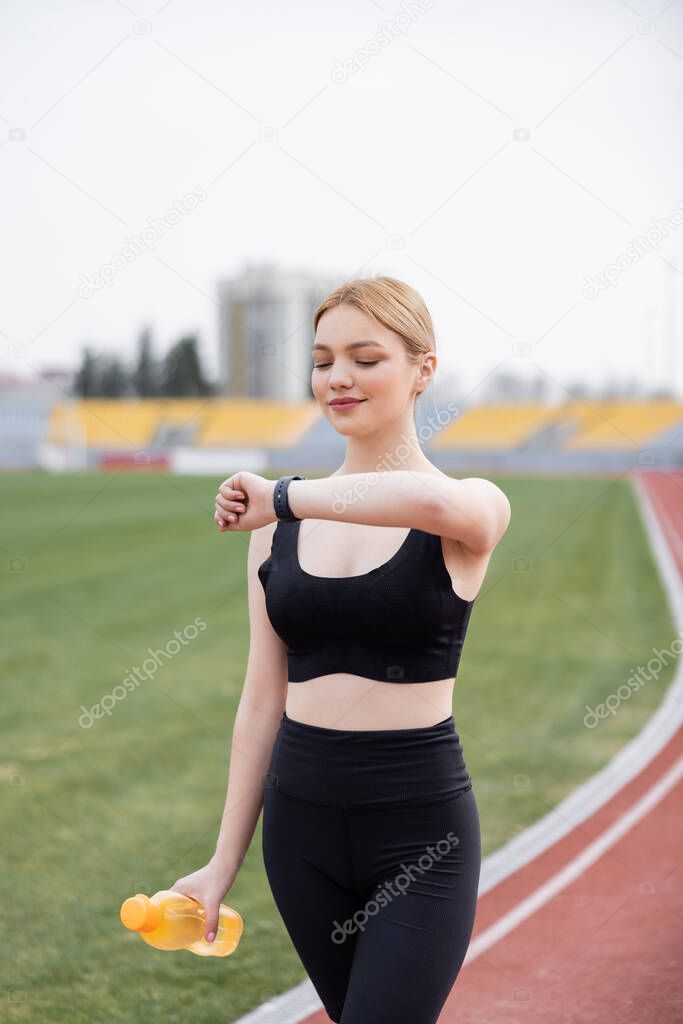young and positive sportswoman looking at fitness tracker on stadium