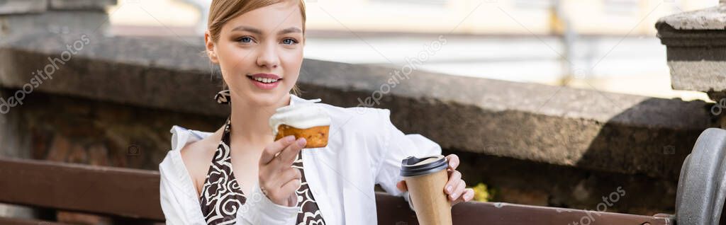 happy young woman with cupcake and takeaway drink looking at camera outdoors, banner