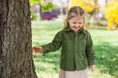joyful girl in green jacket touching tree in park