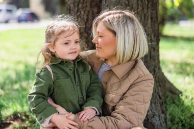 joyful woman looking at daughter while sitting under trees in spring park