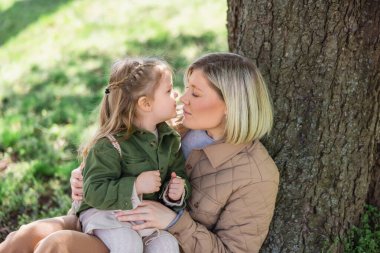 woman and girl in stylish clothes sitting under tree in park and looking at each other