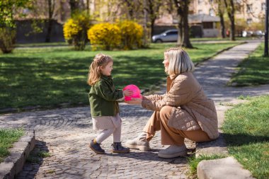 side view of mother and daughter in trendy outfit playing with ball in spring park