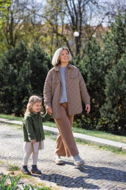 full length of happy and stylish woman with daughter walking in spring park