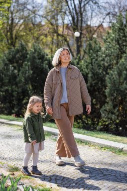 full length of happy and stylish woman with daughter walking in spring park clipart