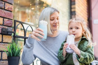 Küçük kız caddedeki kafede gülümseyen annesinin yanında lezzetli bir milkshake içiyor.
