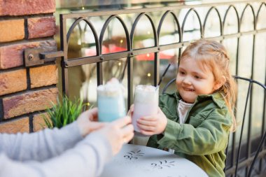 cheerful girl toasting with glasses of milkshake with blurred mom in street cafe clipart