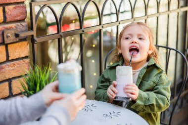 child with open mouth holding glass of milk dessert near blurred mom in street cafe clipart