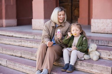 girl blowing soap bubbles near smiling mom and teddy bear on stairs