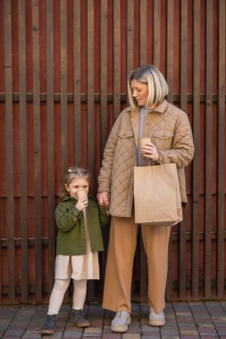 little girl drinking coffee from paper cup near mom with shopping bag