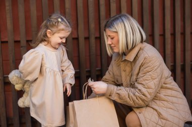 blonde woman holding shopping bags near happy child with teddy bear 