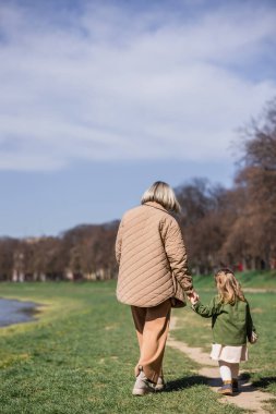 Parkta yürürken el ele tutuşan anne ve küçük kızın arka görüntüsü