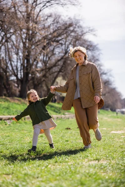 full length view of cheerful mother and daughter holding hands while running in park