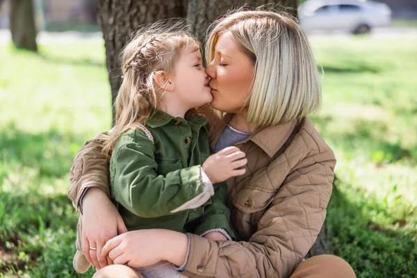 blonde woman kissing little daughter while sitting on green grass in park