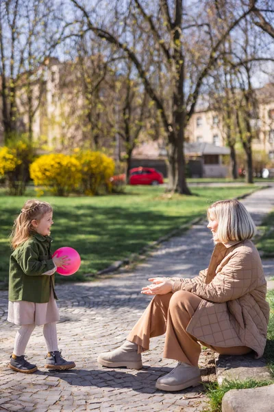 Parkta annesiyle oynarken elinde top tutan neşeli kızın yan görünüşü