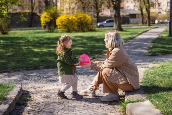 side view of mother and daughter in trendy outfit playing with ball in spring park