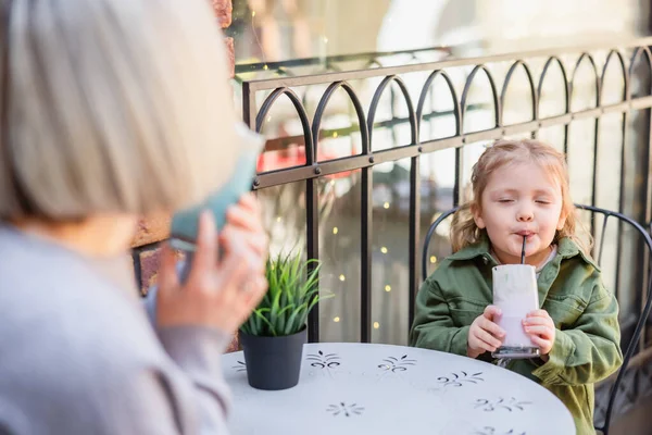 girl with closed eyes enjoying delicious dessert near blurred mom in cafe on street