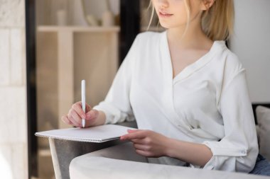 Bluz giymiş genç bir kadının koltuğa defter yazarken çekilmiş görüntüsü. 