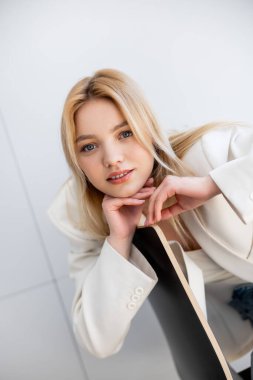 Ceketli genç bir kadın oturma odasındaki sandalyenin yanındaki kameraya bakıyor. 