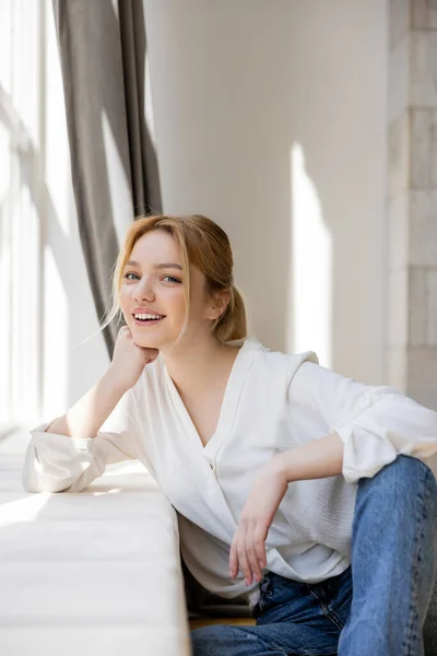 Happy blonde woman in blouse looking at camera near windowsill at home