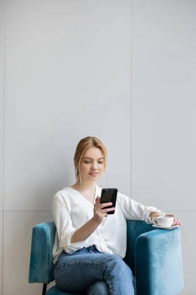 Jovem Mulher Usando Smartphone Segurando Café Poltrona Casa — Fotografia de Stock