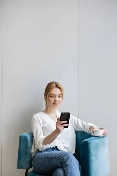 Akıllı telefon kullanan ve evdeki koltukta kahve tutan genç bir kadın.