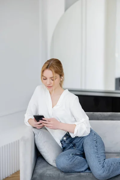 Beyaz bluzlu ve kot pantolonlu sarışın kadın akıllı telefon kullanıyor ve kulaklıkla kanepede müzik dinliyor. 