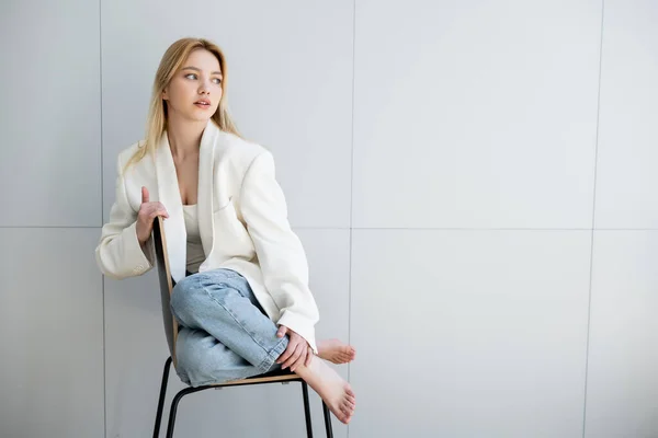 Barefoot woman in jeans and jacket sitting on chair at home