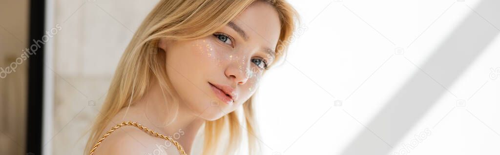 Blonde woman with glitter on cheeks posing at home, banner 