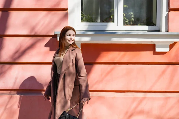 Happy redhead woman in coat standing on street near building — Stock Photo